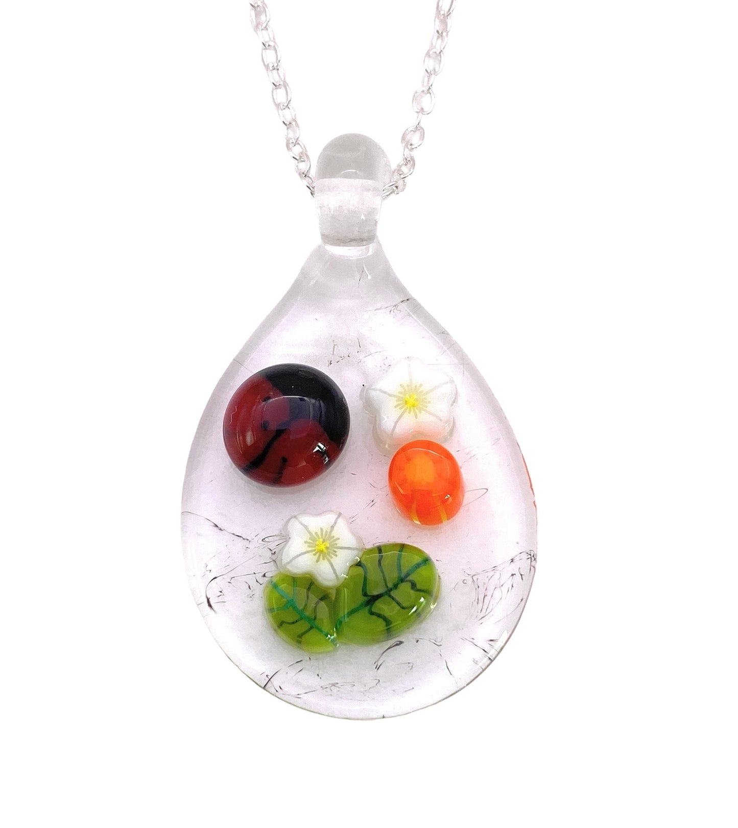 The Handmade Glass Pendant by Calon Glass, known as the Ladybird Pendant, showcases a silver chain adorned with a stunning Millefiori glass teardrop. This exquisite piece contains intricate red and orange orbs, delicate white flowers, and green leaf-like patterns inside, measuring 3cm x 2.5cm.