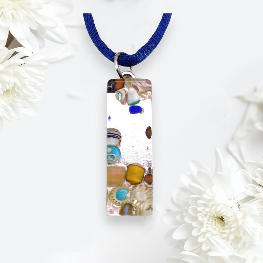 A Murano Passion pendant, crafted with Murano glass set on a silver leaf and measuring 3cm by 1cm, hangs from a blue cord. The piece is strikingly adorned with a mosaic of colorful elements against its silver background. Two white chrysanthemums are elegantly positioned on a light background to highlight the pendant's exquisite beauty and craftsmanship.