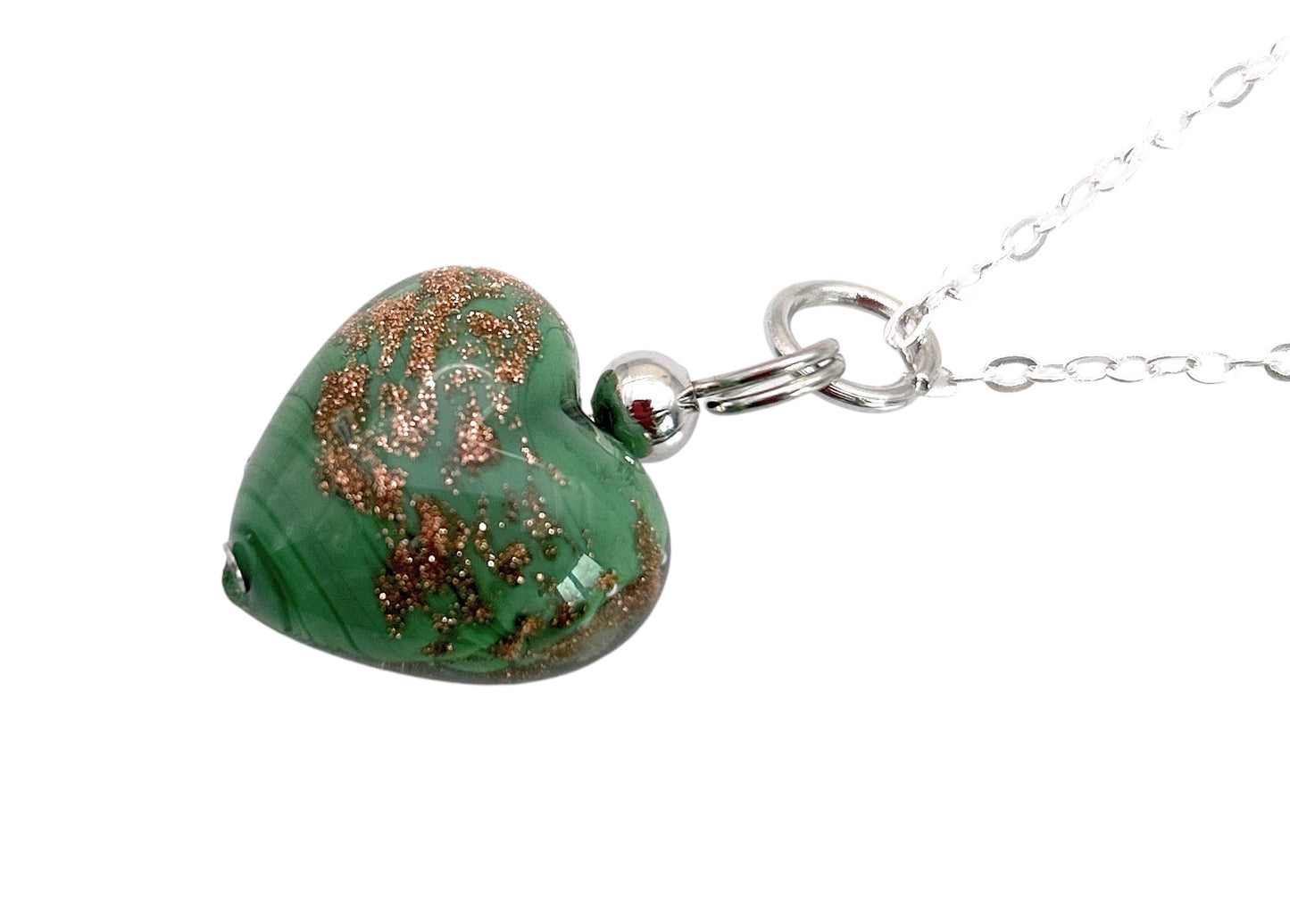 The Murano Passion Heart Pendant is an exquisite piece of jewelry featuring gold glitter accents on a green Murano glass heart suspended from a silver chain, beautifully highlighted against a white background. This genuine craftsmanship comes with a Certificate of Authenticity.