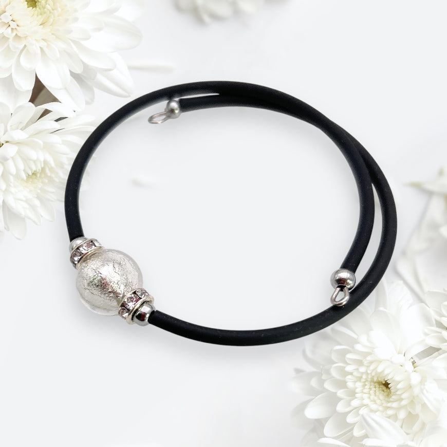 A black leather necklace with a large silver bead and smaller decorative rings rests on a light surface. Surrounding the necklace are white chrysanthemums and elegant accents from Murano Passion's Murano Glass Bead Bracelet, enhancing the image's allure.