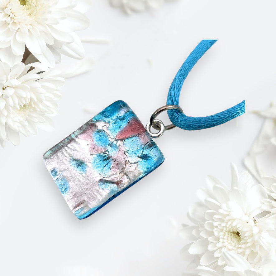 The Murano Glass Pendant by Murano Passion, named the Millefiori Flowers on Silver Leaf, is a dainty piece measuring 2cm x 1.5cm. It hangs from a blue cord and is surrounded by white chrysanthemums on a light backdrop, showcasing abstract patterns in blue, pink, and silver hues.