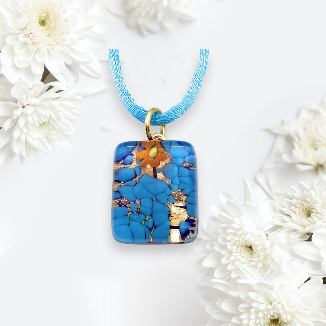 The Murano Passion's Murano Glass Pendant in Blue on Gold Leaf, sized at a dainty 2cm x 1.5cm (MGPP28), hangs from a blue chain amidst white flowers on a light background.