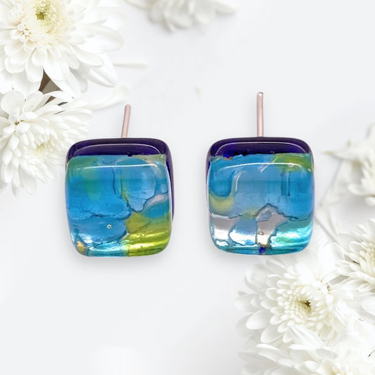 Close-up of Murano Passion's Murano Glass Earrings - Multi/Silver, Dainty 8mm Diameter (MGE19) featuring a vibrant abstract design in blue, green, and silver. The background has soft white flowers, enhancing these genuine handmade accessories' elegance from Venice.