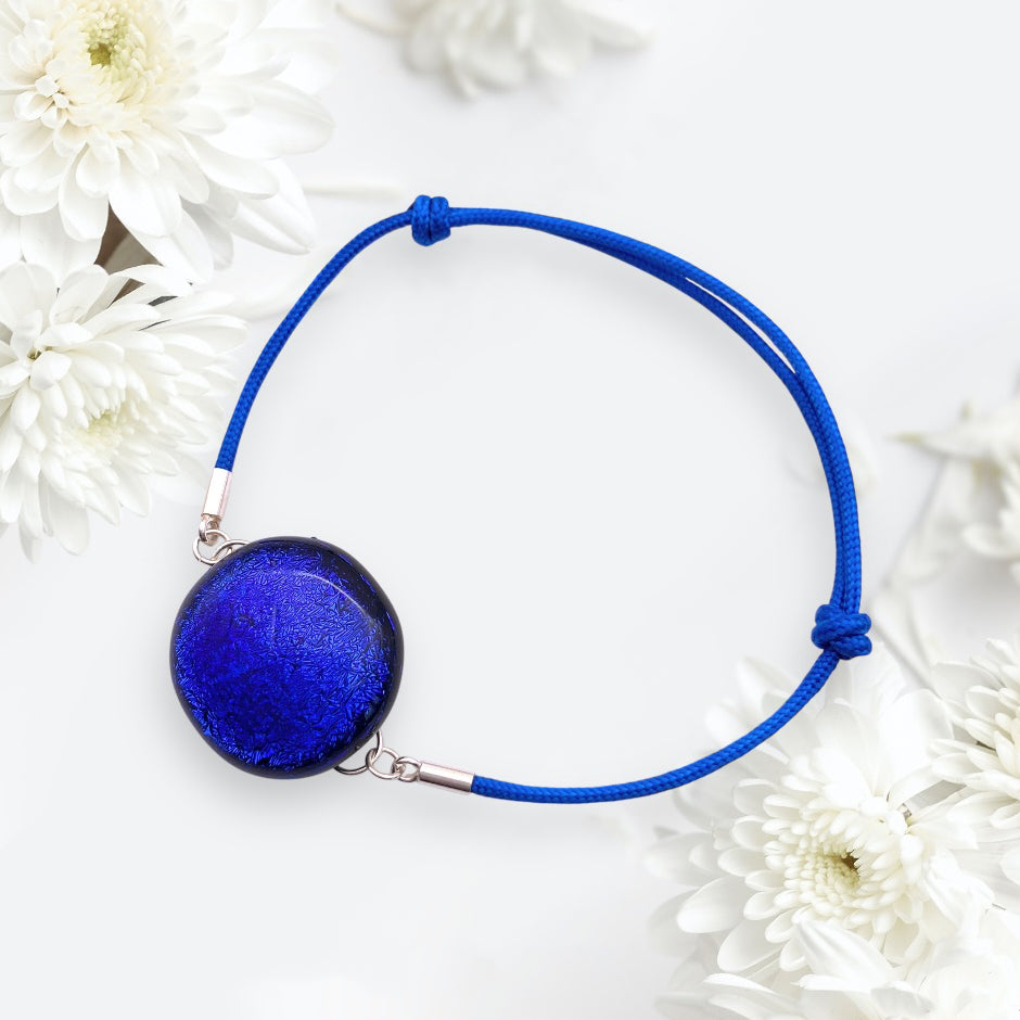 The Calon Glass handmade dichroic glass bracelet (CGBR4) in vibrant blue is elegantly showcased against white flowers. It features a circular, reflective pendant on a securely tied thin cord.