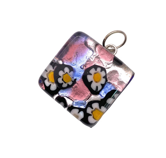 This Murano Passion pendant features a square design made from Murano glass, showcasing Millefiori flowers with white petals and yellow centers set against a black backdrop. Elegant pink and purple tones shimmer on the surface, while a small metal loop ensures easy attachment to any necklace or chain. The pendant measures 2cm by 2cm and comes with a certificate of authenticity.