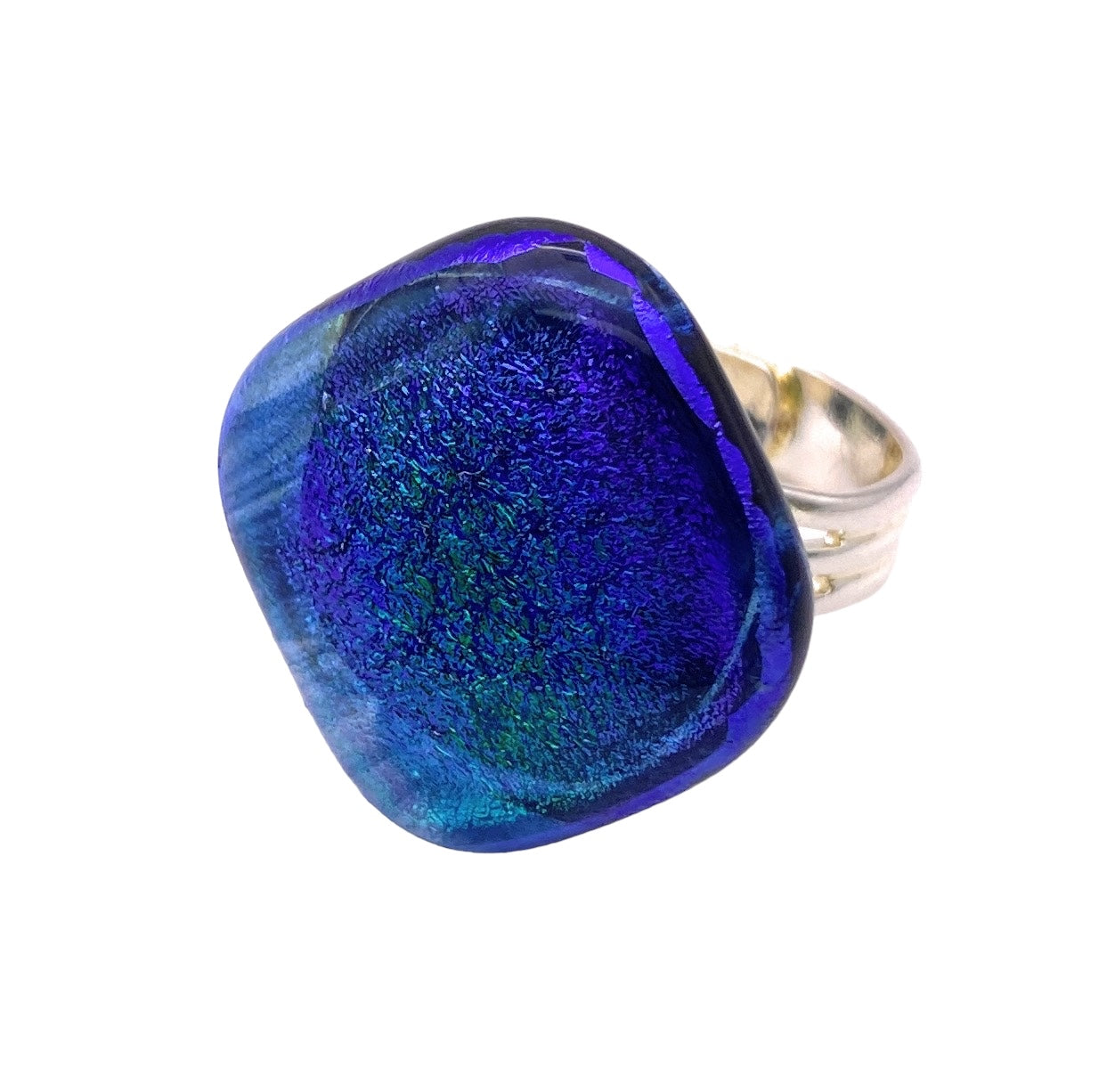 Introducing the Calon Glass Handmade Dichroic Glass Ring, an exquisite piece of jewelry featuring a large, square-shaped dichroic glass stone. This adjustable ring shimmers with iridescent blue and purple tones, reminiscent of deep ocean hues accented by hints of green. A modern and artistic creation, this 2cm textured silver ring is a stunning example of handmade glass artistry.