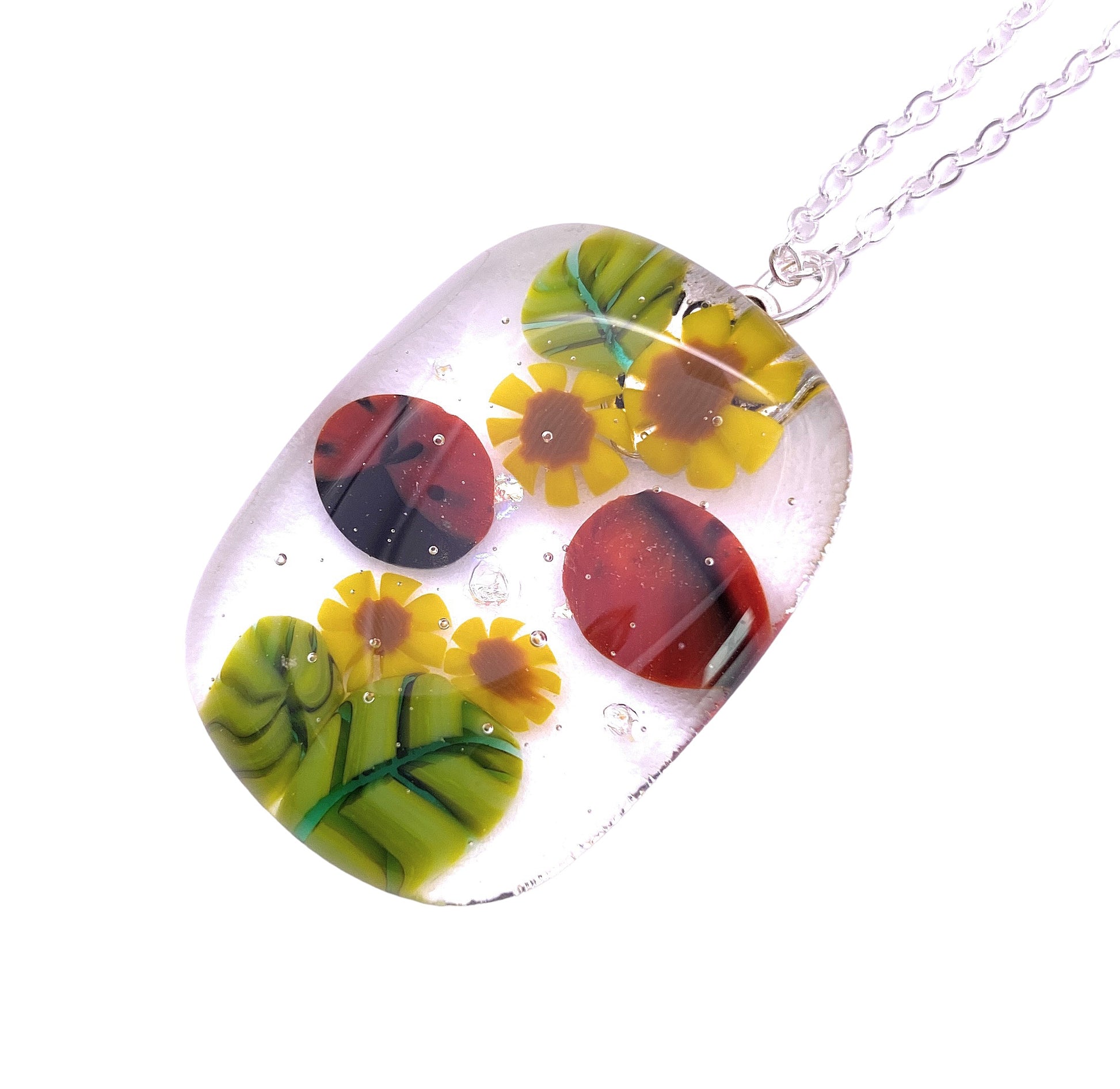 The Calon Glass Ladybird Pendant is a 3.5cm x 2.5cm handmade glass pendant featuring vibrant yellow flowers, red circles, and green leaves encapsulated in clear resin on a silver chain for a unique touch.