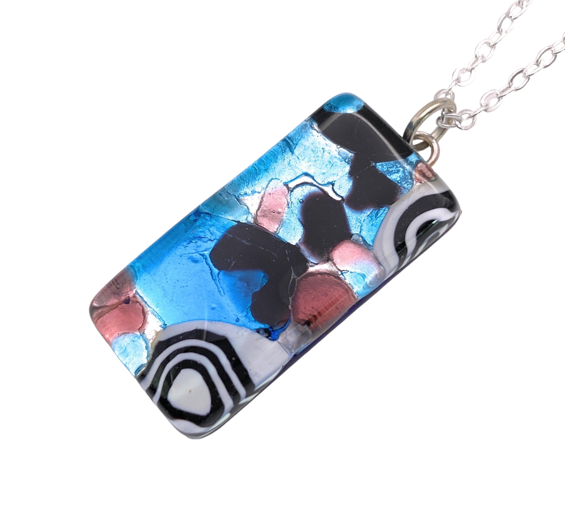 A Murano Passion pendant, featuring a rectangular Murano glass design accented with abstract patterns in blue, black, pink, and white, elegantly hangs from a silver chain. The Millefiori pendant on silver leaf measures 3cm x 1.5cm (MGPL2) and showcases layered shapes and swirling lines for a vibrant and artistic appearance. This exquisite piece of Murano Glass Jewellery is accompanied by a Certificate of Authenticity.