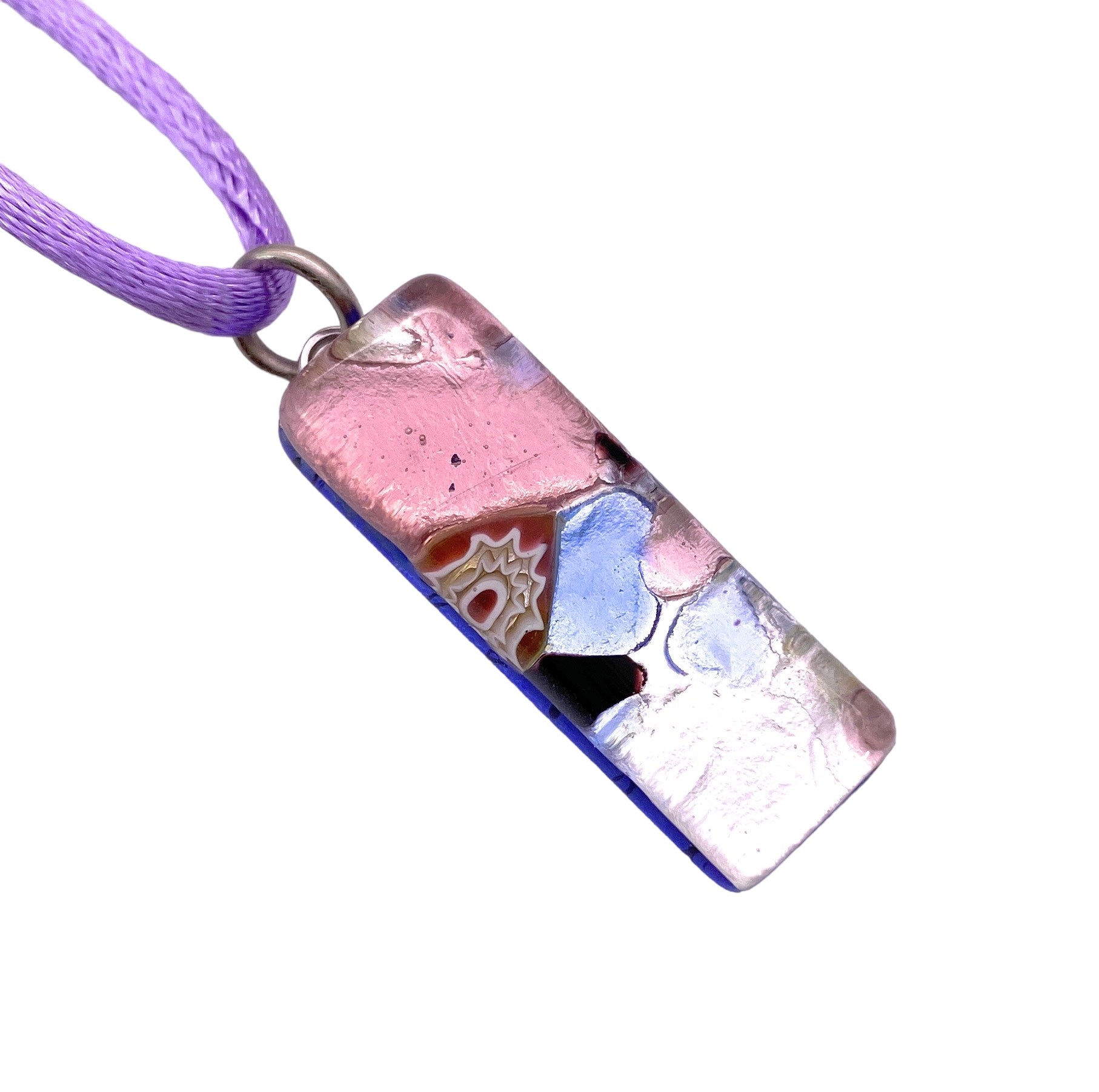 The Murano Passion Glass Pendant on Silver Leaf, measuring 3cm x 1cm (MGPL15), boasts a rectangular design paired with a purple cord. It beautifully blends pink, blue, and silver hues, accented by a small geometric flower pattern, all finished in glossy glass that exemplifies the sophistication of Murano Glass Jewellery.