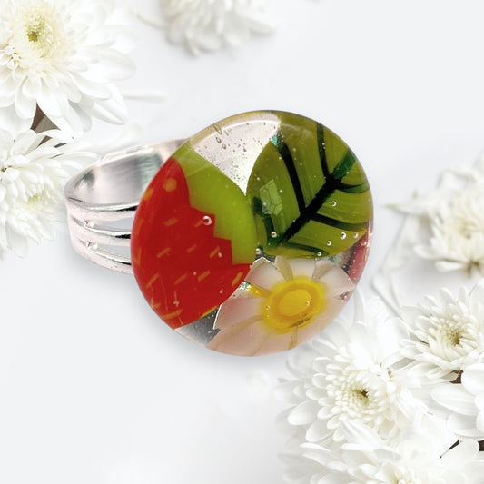 Calon Glass's Strawberry Ring (CGR7) is a handmade glass ring featuring a silver band and a round, colorful enamel top with a strawberry, green leaf, and white flower. The background showcases Millefiori-inspired patterns reminiscent of white chrysanthemum flowers.