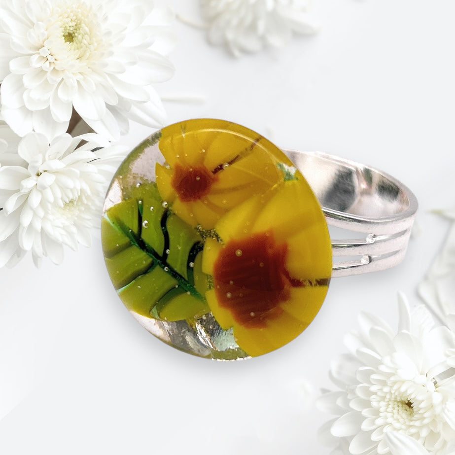 The Sunflower Ring by Calon Glass features a silver band adorned with a circular handmade glass design, embellished with vibrant yellow and red flowers complemented by lush green leaves. Framed by white chrysanthemums on a delicately blurred background, this Flower Ring beautifully captures the intricate artistry of Millefiori pieces.