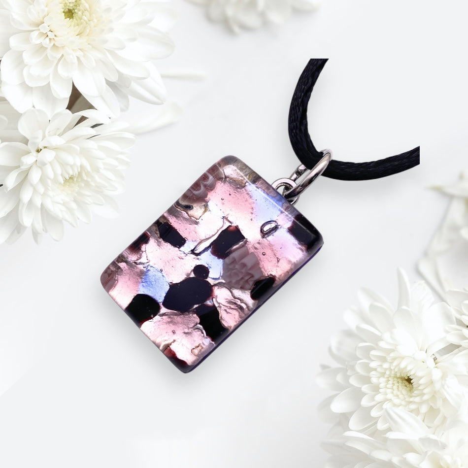 The Murano Passion Pendant, made with Silver Leaf, features a 2.5cm x 1.5cm rectangular design with abstract pink, black, and white patterns suspended from a black cord necklace. It is accompanied by delicate white chrysanthemum flowers on a light background and includes a Certificate of Authenticity.