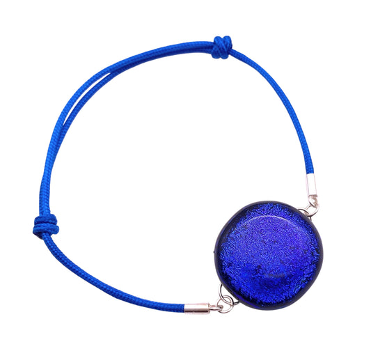 The Calon Glass bracelet features a 2cm round, deep blue dichroic glass pendant on a sleek, adjustable blue cord with metallic clasps, creating a striking and elegant accessory.