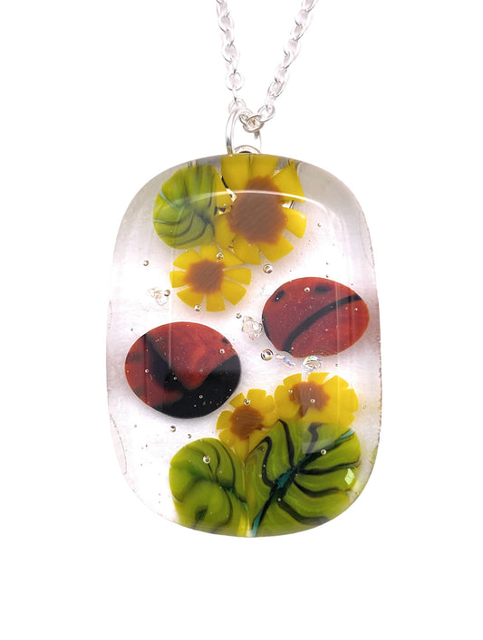 The Handmade Glass Pendant by Calon Glass, known as the Ladybird Pendant or Flower Floral Pendant, measures 3.5cm x 2.5cm and features vibrant floral designs in yellow, green, and red sealed in clear resin. This handmade piece is rectangular with rounded edges and contains air bubbles.