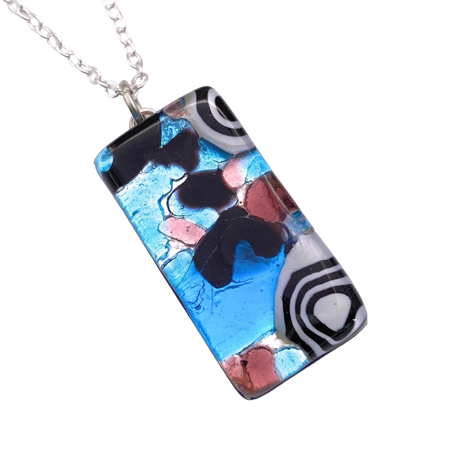 Presenting the Murano Passion Murano Glass Pendant, Millefiori Pendant on Silver Leaf (3cm x 1.5cm, MGPL2), adorned with a silver chain. This rectangular piece showcases an abstract design featuring swirling patterns in vibrant blue, black, pink, and white hues that create a unique artistic appearance. Accompanied by a Certificate of Authenticity, this pendant is truly special.