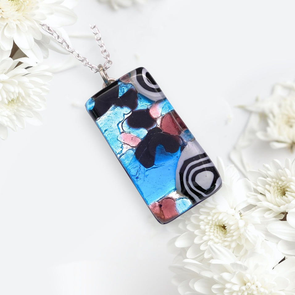 The Murano Passion MGPL2 Pendant is a rectangular Murano Glass creation showcasing an abstract design in shades of blue, black, pink, and white. It hangs elegantly on a silver chain and is complemented by delicate white flowers against a light background. Complete with a Certificate of Authenticity, this Millefiori Pendant on Silver Leaf (3cm x 1.5cm) is an ideal accessory for any occasion.