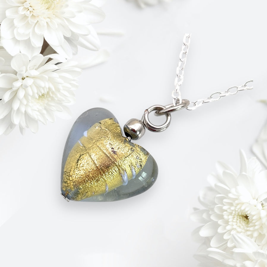 A Murano Passion heart-shaped pendant (MHT13), crafted from blown Murano glass with a gold leaf design, hangs elegantly from a silver chain, surrounded by white daisies on a light background.