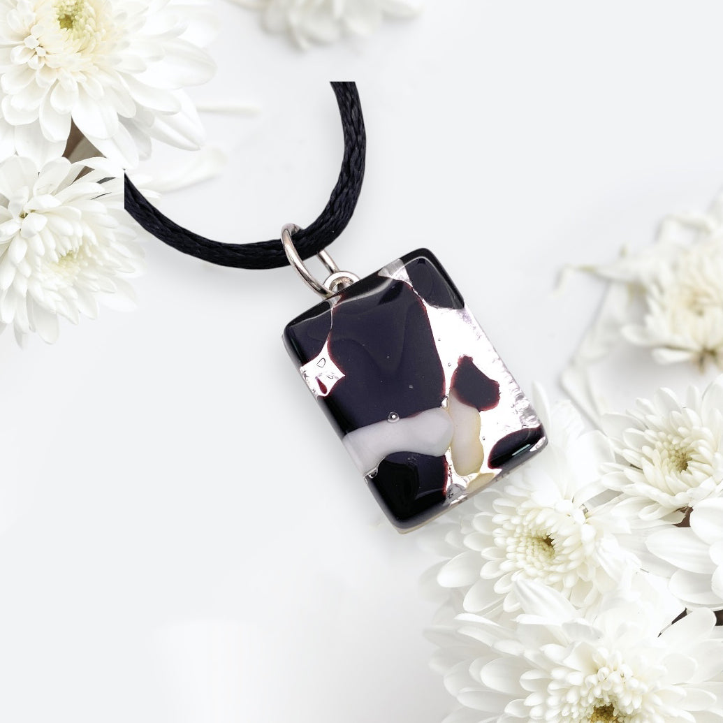 A rectangular pendant from Murano Passion hangs gracefully from a black cord, showcasing a blend of black on silver leaf with a smooth, glossy finish. It's accompanied by clusters of white chrysanthemum flowers on a light background, evoking the exquisite craftsmanship found in the Murano Glass Pendant collections. This dainty piece measures 2cm x 1.5cm and is designated as MGPP7.