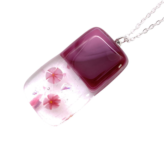 The Calon Glass Handmade Dichroic Glass Pendant is an elegant rectangular necklace with a smooth, glossy surface crafted from handmade glass. The top half showcases a solid deep pink hue, while the bottom half is clear and features embedded Millefiori pink flowers accented with small iridescent details. This beautiful piece is suspended gracefully from a delicate silver chain and measures 4cm x 2cm.