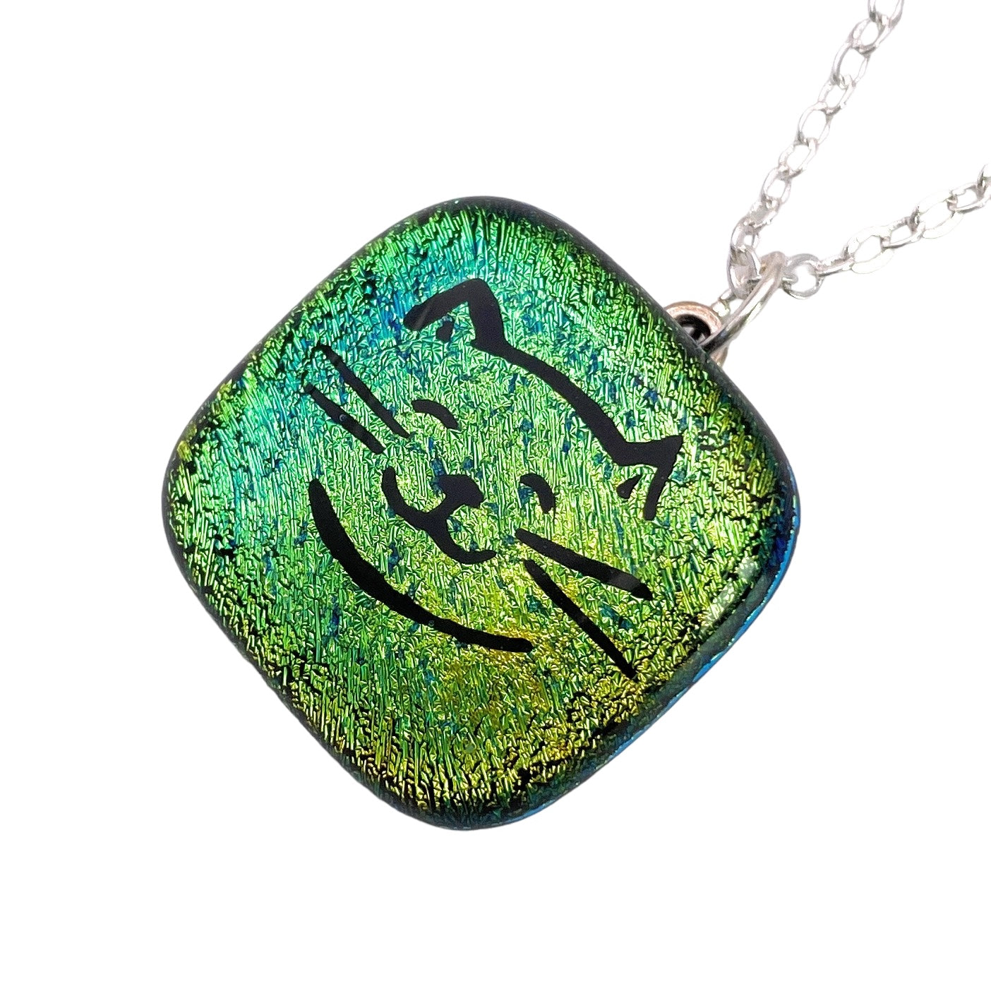 The Handmade Dichroic Glass Pendant by Calon Glass is a striking square piece, measuring 2cm x 2cm. It features an iridescent finish in green and yellow with a simple black outline of a smiling cat face and is elegantly attached to a silver chain necklace.