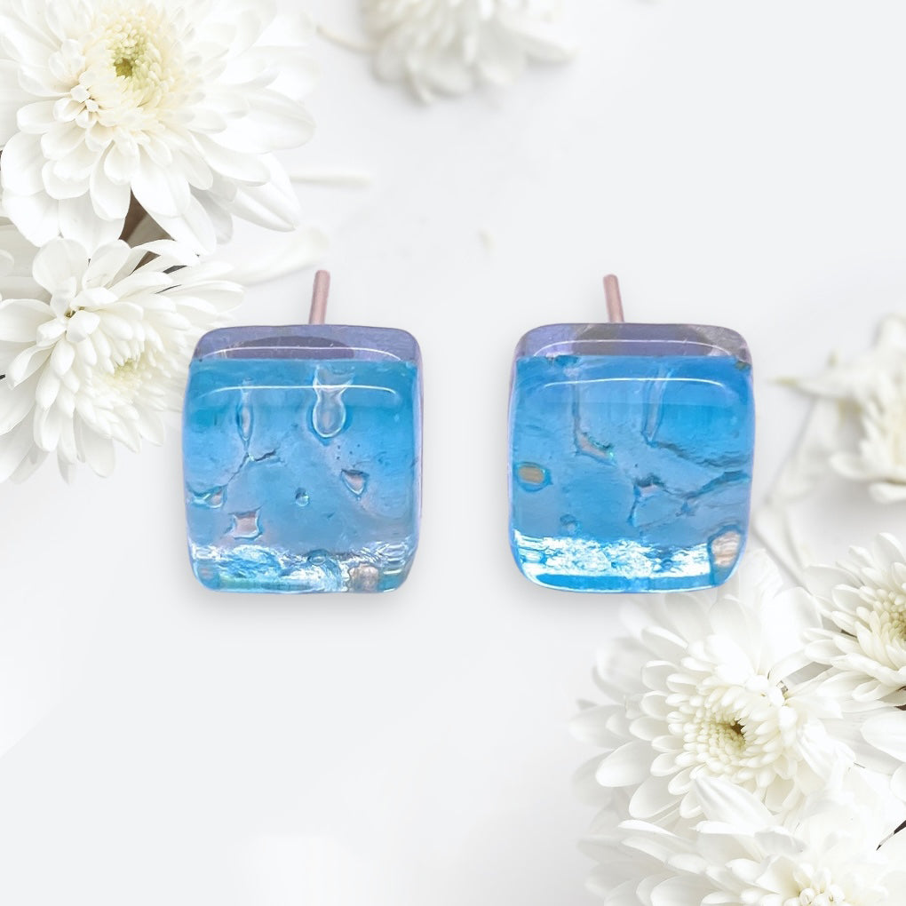 The Murano Passion earrings, titled "Murano Glass Earrings - Blue/Silver, Dainty 8mm Diameter (MGE18)," evoke the artistry of Murano Glass with their square blue design. Displayed among white chrysanthemums on a gentle, light backdrop, these handmade earrings feature a translucent texture that beautifully complements the surrounding delicate flowers.