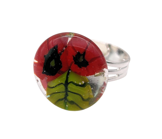 The Poppy Ring by Calon Glass features a round, transparent glass setting that beautifully displays a vibrant red poppy design, complete with black centers and green leaves. The intricate Millefiori flower pattern adds an exquisite charm to this handmade glass ring.