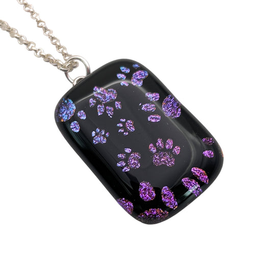 Introducing the Handmade Dichroic Glass 'Paw Print' Pendant by Calon Glass, featuring a rectangular design measuring 3cm x 2cm. This exquisite piece boasts shiny purple paw prints on a black pendant and is elegantly suspended from a silver chain.