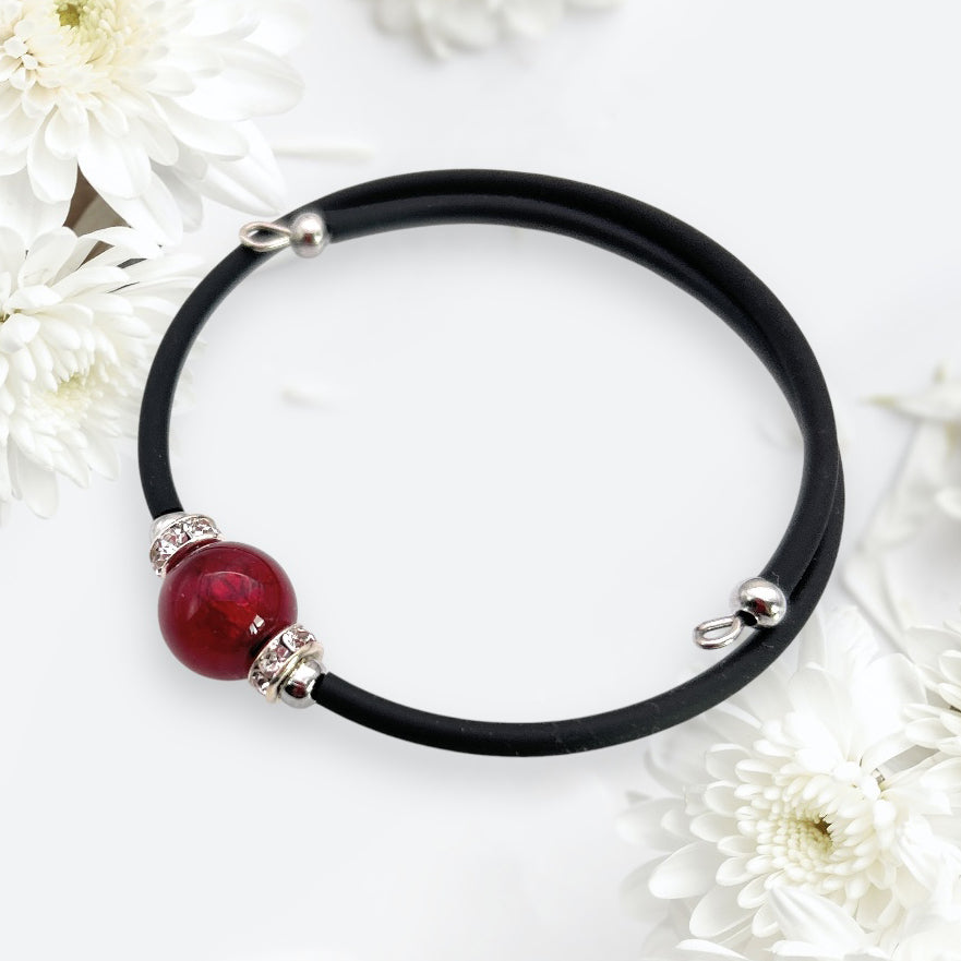 A captivating Murano Passion memory wire bracelet, showcasing an exquisite 1cm Murano glass red bead with silver accents, lies gracefully on a white background, encircled by delicate white flowers.