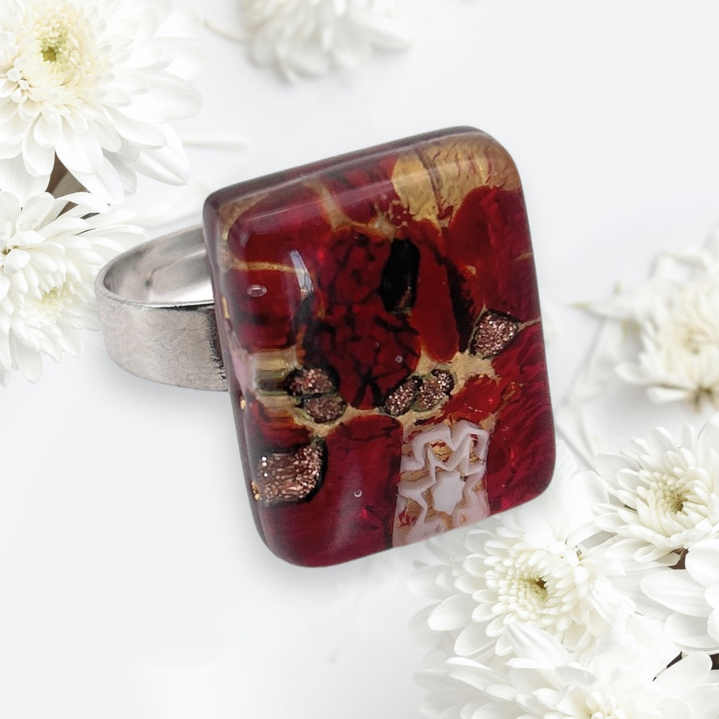 A Murano Passion Millefiori Flower on Gold Leaf glass ring, measuring 2cm x 1.5cm, is elegantly displayed against a backdrop of white flowers. The vibrant patterns of this authentic Murano Glass piece beautifully contrast with the delicate petals.