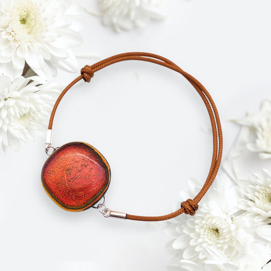 A handmade Glass Bracelet from Calon Glass, featuring a burnt orange dichroic glass centerpiece, is surrounded by white chrysanthemum flowers on a light background.