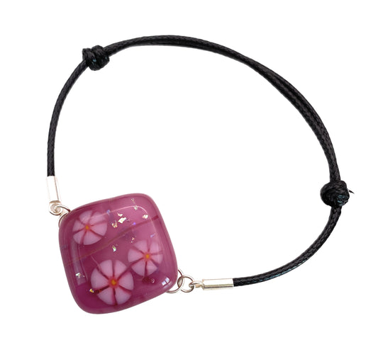 The Calon Glass Handmade Dichroic Glass Bracelet (CGBR1) features a square, purple bead with pink and white millefiori flowers, set on a black cord with silver clasps.