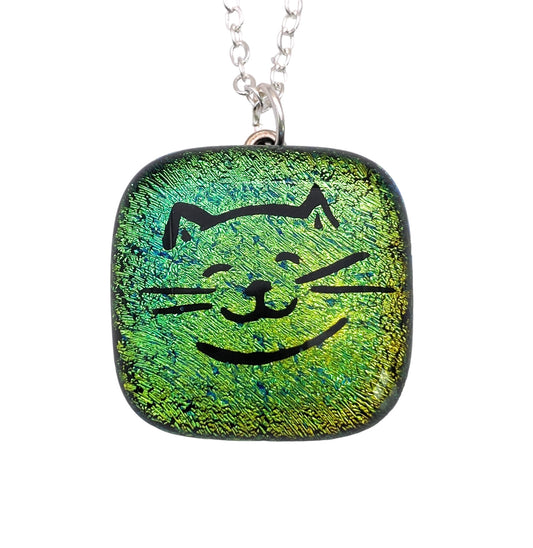 The Handmade Dichroic Glass Pendant by Calon Glass features a square design measuring 2cm x 2cm, showcasing a green textured surface with an adorable smiling cat face. This charming piece hangs elegantly from a silver chain.