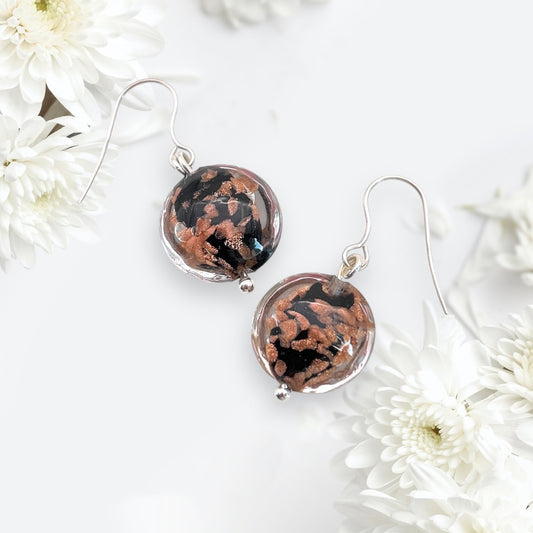 The Murano Passion Murano Glass Beaded Earrings, featuring black and copper swirls on 925 sterling silver dropper hooks, are artistically presented among delicate white flowers on a soft white background, reflecting the opulent Venetian heritage.