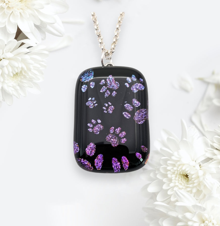 The breathtaking Calon Glass necklace features a handmade dichroic glass 'Paw Print' pendant, measuring 3cm by 2cm. The black rectangular pendant is decorated with vibrant paw prints and delicately set against a white backdrop with lovely white flowers. It hangs gracefully from a silver chain.