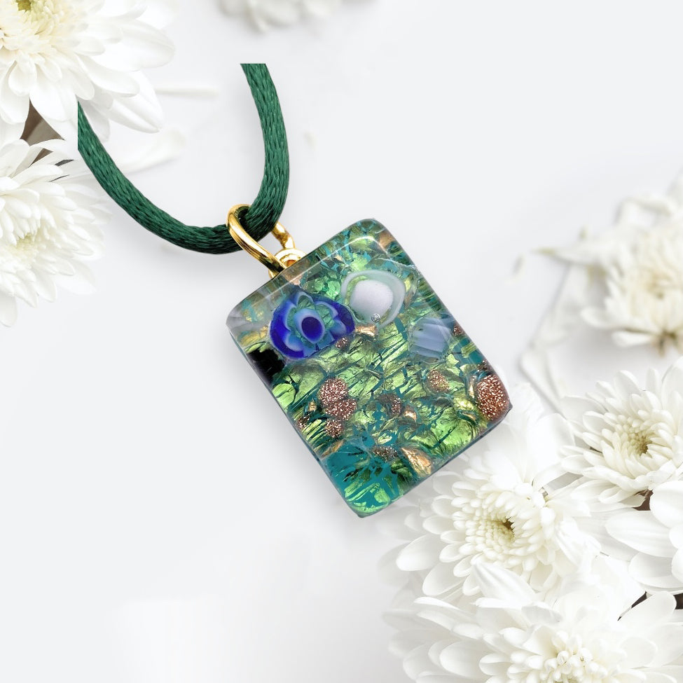 A dainty rectangular 2cm x 1.5cm Murano Passion glass pendant with a green rope necklace, features blue and white floral designs with gold accents. Elegantly set on a white background surrounded by chrysanthemums, it embodies the timeless charm of authentic Murano jewellery (MGPP34).