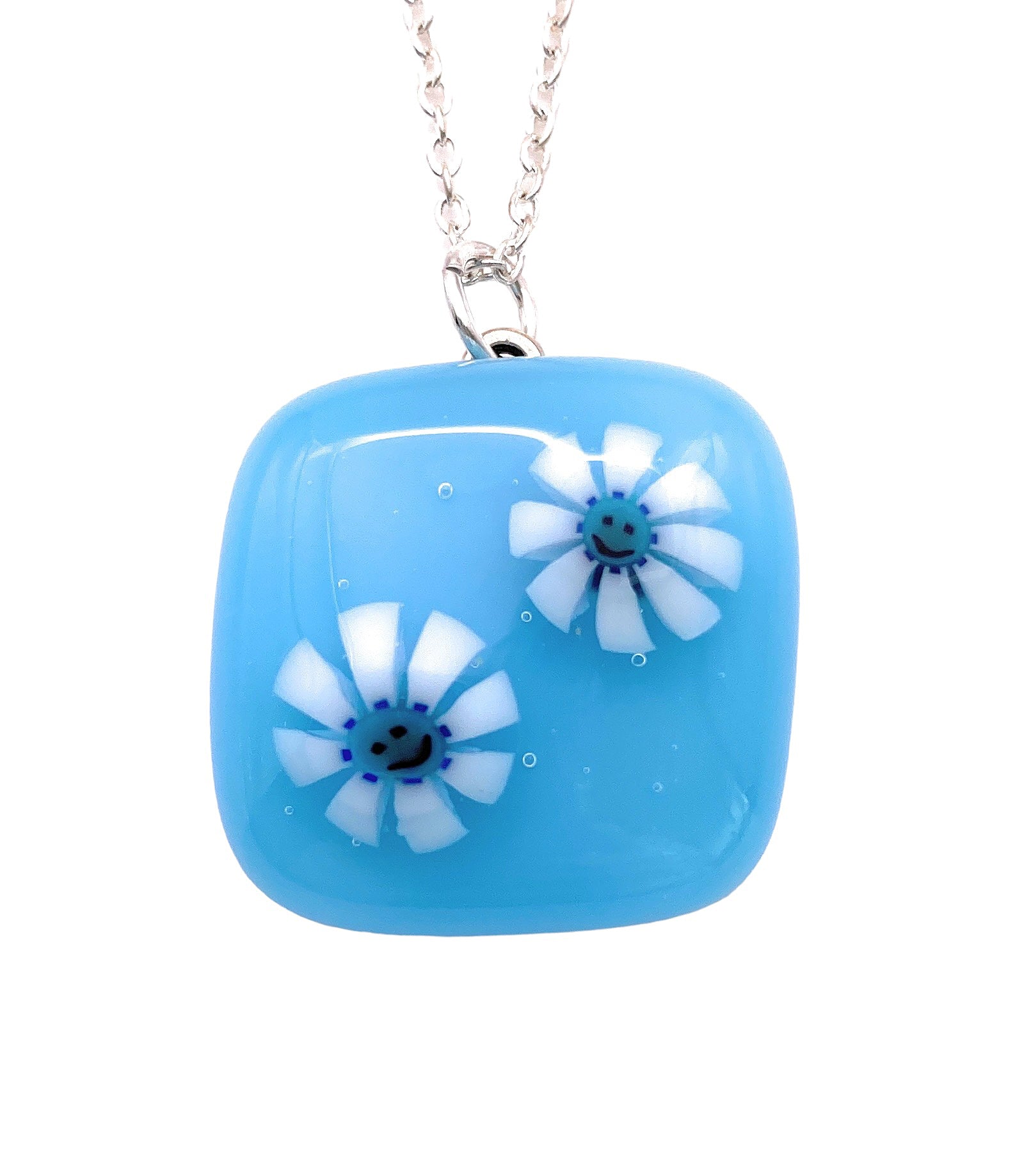 The Calon Glass Handmade Smiley Face Millefiori Flower Pendant showcases a sky-blue square with rounded edges, adorned with two daisy-like flowers in white, featuring blue centers and smiley faces. This elegant piece measures 2cm x 2cm and is gracefully suspended on a silver chain.