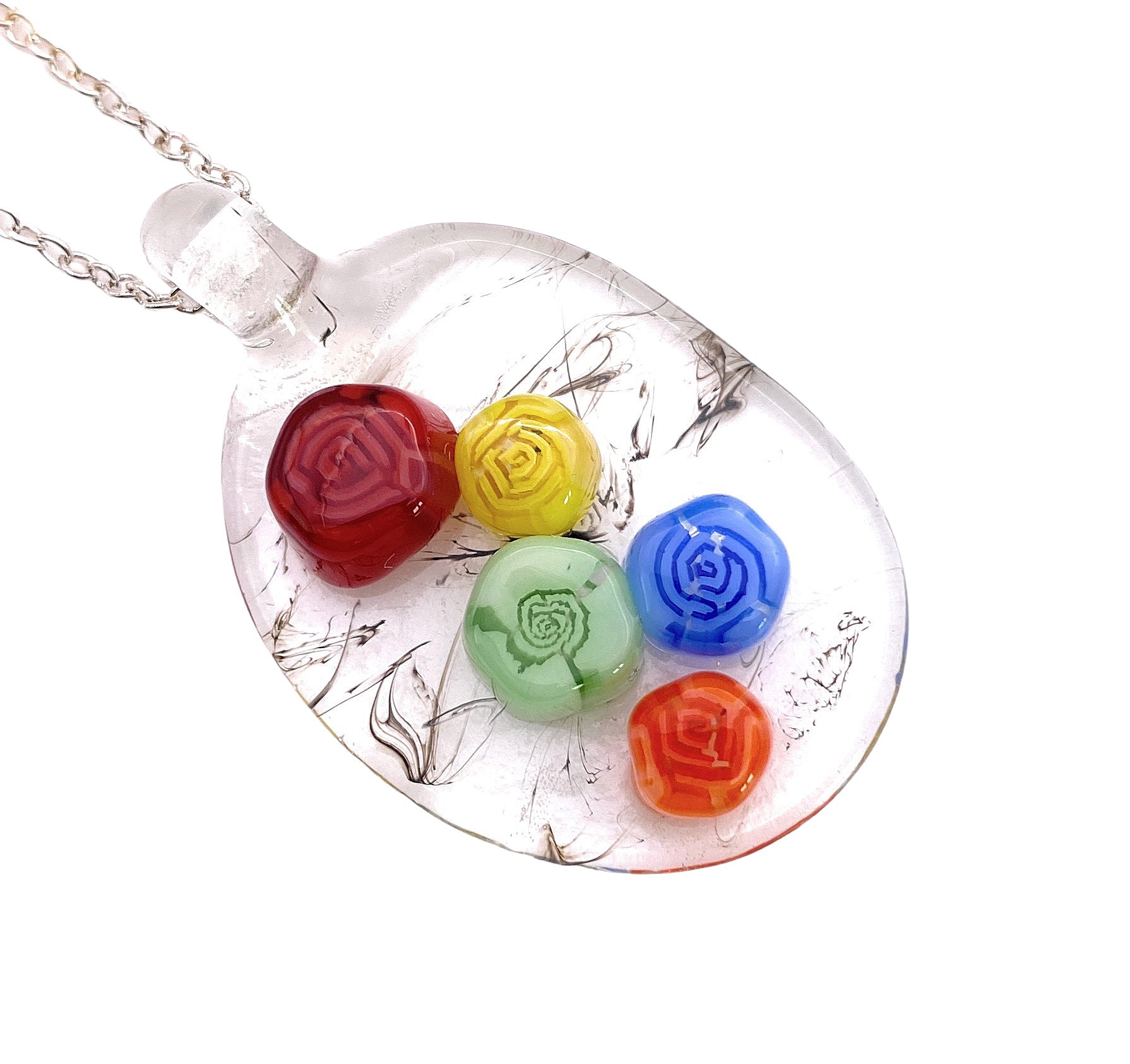 The Calon Glass Flower Pendant is a handcrafted piece featuring a transparent oval design adorned with colorful rose-shaped patterns in red, yellow, green, blue, and orange. Made from Millefiori glass, it gracefully hangs from a silver chain and is set against a white backdrop. The pendant measures 3cm x 2cm.