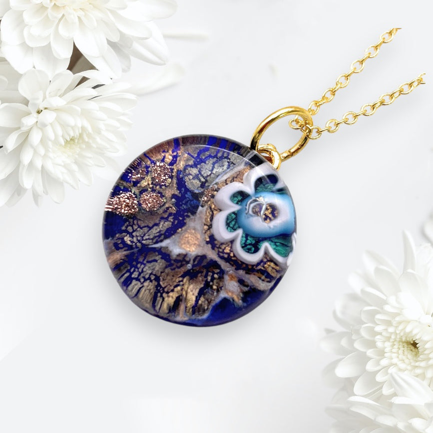 Close-up of the Murano Passion Murano Glass Round Pendant made with Millefiori Flower - 2cm (MGPV20), showcasing a captivating blue and gold abstract pattern along with a decorative floral design. The pendant gracefully hangs from a gold chain, surrounded by white flowers against a light background. It includes a Certificate of Authenticity to guarantee its exquisite craftsmanship.