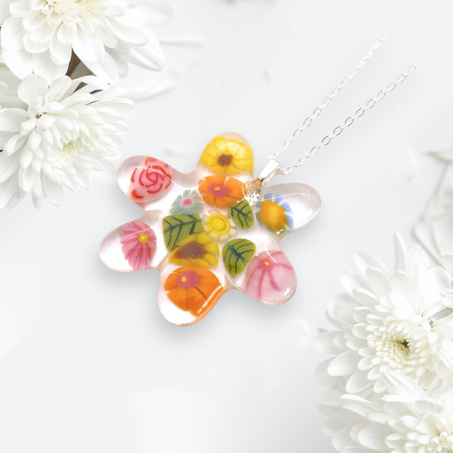 A flower-shaped pendant crafted from Millefiori glass by Calon Glass hangs gracefully from a silver chain, surrounded by white chrysanthemums. This Handmade Glass Pendant, known as the Millefiori Flower Pendant (CGP31), measures a chunky 4.5cm x 4cm and showcases vibrant designs in red, yellow, pink, and orange with green leaves, capturing the essence of handmade art.