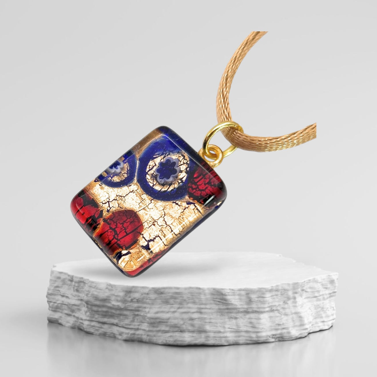 A vibrant Murano Glass Pendant from Murano Passion, featuring contemporary Millefiori patterns with striking blue, red, and gold accents on a gold leaf base, is elegantly showcased on a textured stone platform against a plain backdrop. The dainty 2cm x 1.5cm pendant (MGPP27) hangs gracefully from a gold chain.