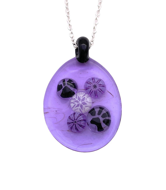 A pendant from Calon Glass, featuring a purple teardrop shape crafted from handmade glass, showcases abstract black and white circular patterns. This pendant, measuring 3cm x 2.5cm, hangs elegantly from a silver chain and boasts varying designs for a dynamic and artistic appearance.