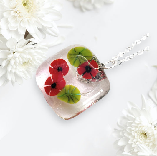 Introducing the Calon Glass Poppy Pendant (CGP29), a stunning handmade glass creation. This exquisite flower pendant showcases a square charm adorned with vibrant red poppy flowers and lush green leaves, beautifully contrasted against a backdrop of delicate white chrysanthemum flowers, offering an elegant fusion of colors and textures.