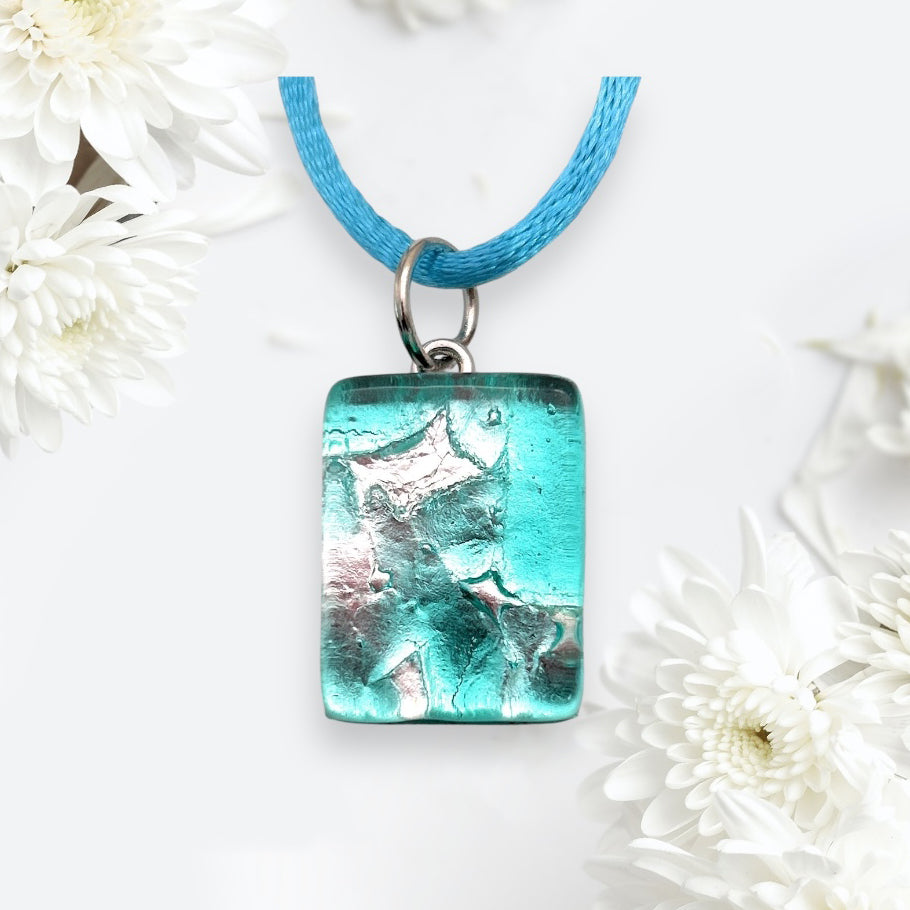 A Murano Passion turquoise and silver rectangular millefiori pendant from the collection of Murano Glass Pendant made using Silver Leaf (MGPP20), delicately designed on a blue cord, is beautifully surrounded by white chrysanthemums on a light background. The pendant showcases a textured surface with metallic accents and vivid turquoise hues, evoking the timeless artistry of Venetian Murano jewelry.