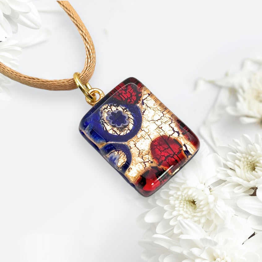 The Murano Passion Millefiori Pendant on Gold Leaf, measuring a dainty 2cm x 1.5cm (MGPP27), features a captivating fusion of multicolored gold, blue, and red patterns. It elegantly hangs from a beige cord and is set against a white background embellished with delicate white flowers, encapsulating the essence of Murano Glass Jewellery.