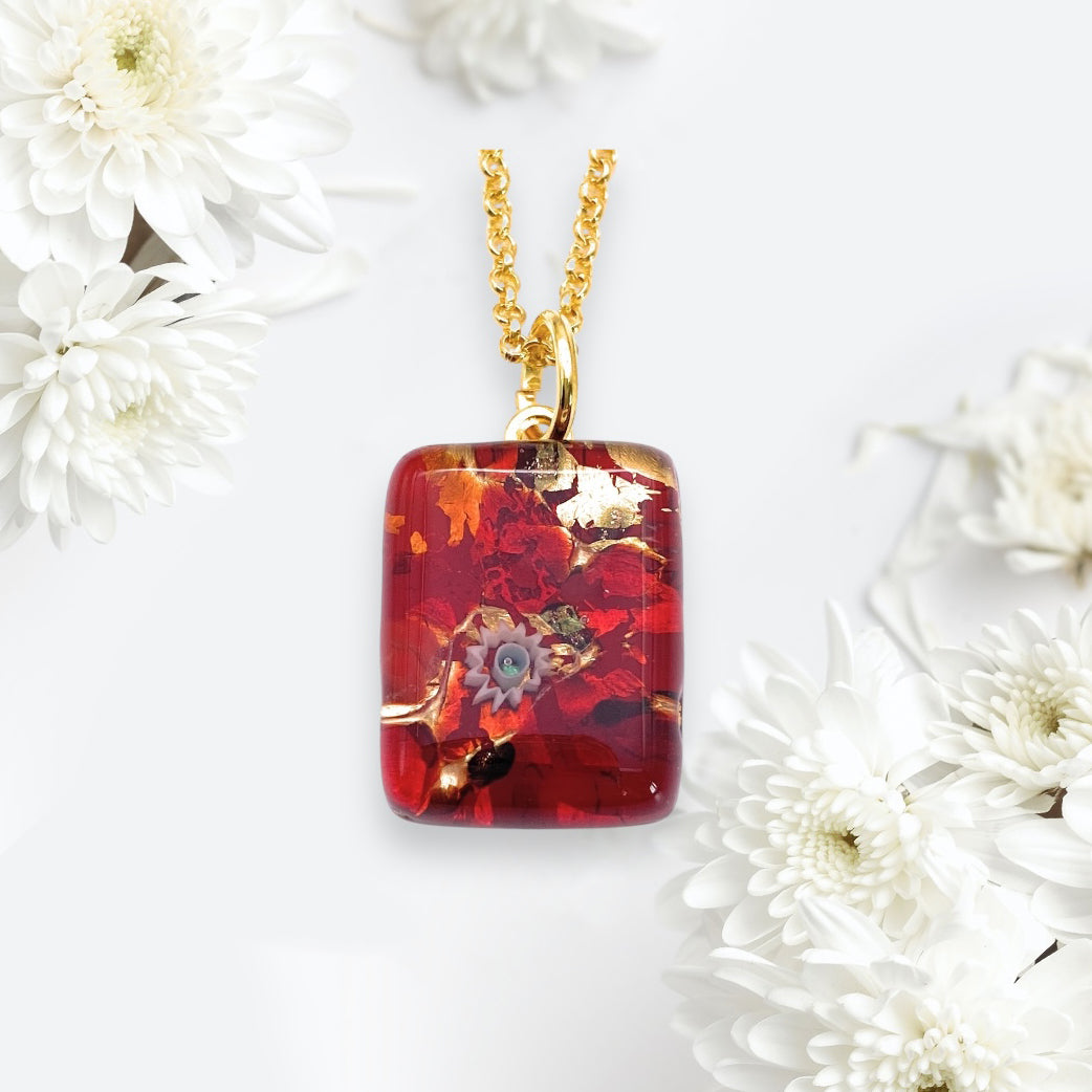 A rectangular Murano Glass Pendant, Multicolour on Gold Leaf by Murano Passion, measuring 2cm x 1.5cm, hangs from a gold chain, surrounded by white chrysanthemums on a light background.