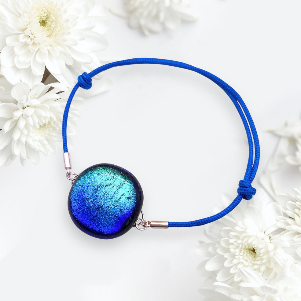 The Glass Bracelet by Calon Glass (CGBR2) features a blue cord with a dichroic glass centerpiece in vibrant blue and black, elegantly surrounded by Millefiori flowers.