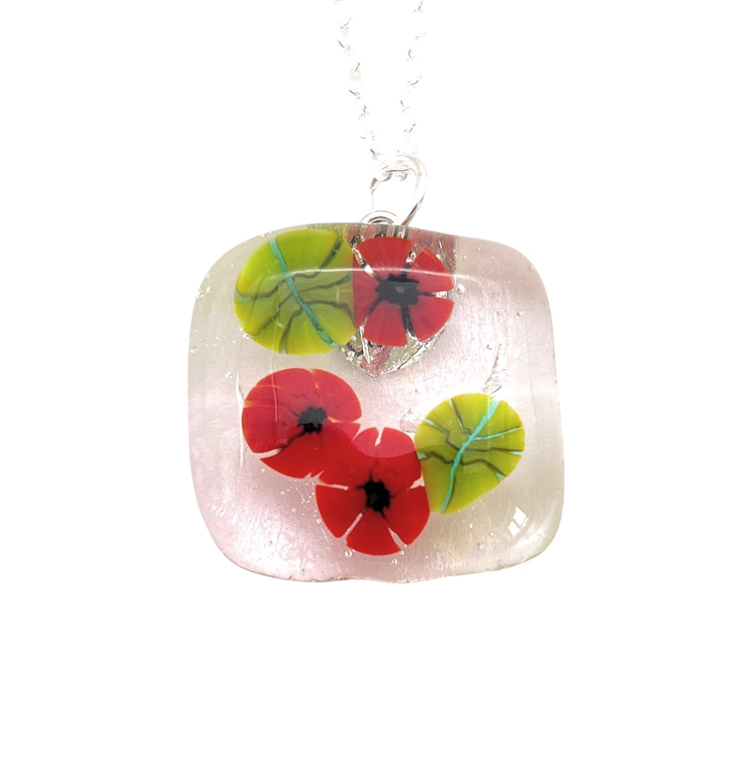 The Poppy Pendant by Calon Glass, identified as CGP29, is a handmade glass pendant that features vibrant red flowers and green leaves set within its square-shaped transparent frame. Crafted from dichroic glass using kiln-fusion techniques, this striking flower pendant hangs gracefully from a chain, adding an artistic flair to any outfit.