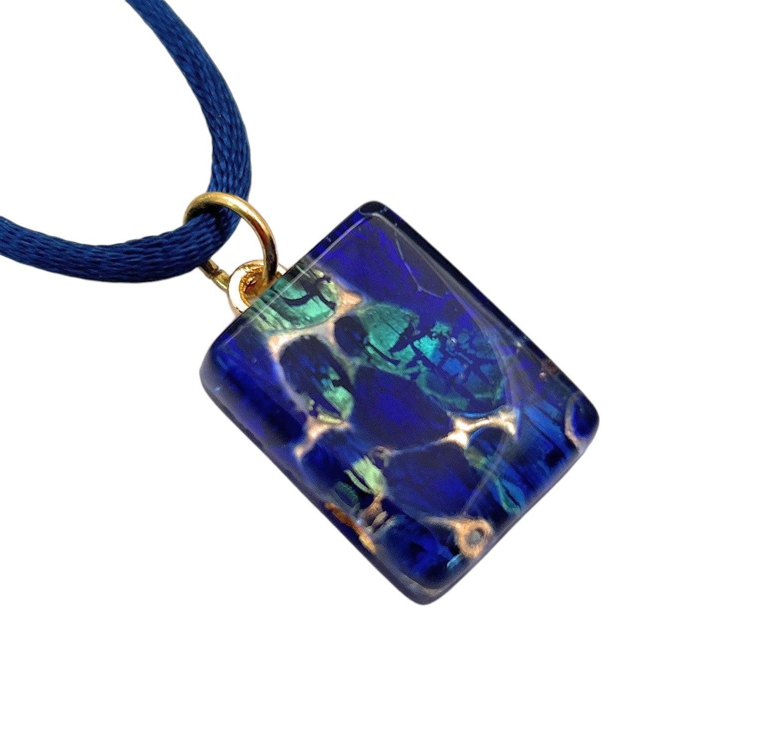 The Murano Passion pendant, named "Murano Glass Pendant, Navy on Gold Leaf - Dainty 2cm x 1.5cm (MGPP16)," features a rectangular glass piece with a captivating blue and green iridescent pattern. The surface of the pendant shimmers as it reflects light. A blue cord necklace is elegantly looped through a small gold ring at the top, highlighting its authentic craftsmanship.