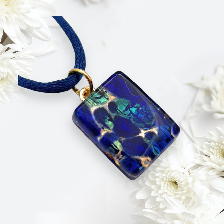 The Murano Passion Murano Glass Pendant in Navy on Gold Leaf, measuring a dainty 2cm x 1.5cm (MGPP16), features a vibrant blue rectangular design with gold accents, elegantly hanging from a dark blue cord. This exquisite piece is set against delicate white flowers on a light background and comes complete with a Certificate of Authenticity to guarantee its elegance and craftsmanship.