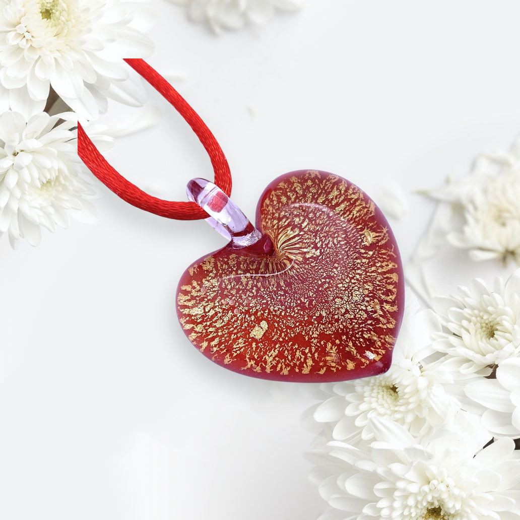 A stunning heart pendant crafted from Murano glass, featuring intricate gold patterns, hangs elegantly from a red cord. It is beautifully displayed among white chrysanthemums on a light background. Each piece of Murano Passion’s Murano Glass Heart Pendant - 3cm (MHT11), comes with a Certificate of Authenticity to guarantee its genuine craftsmanship.